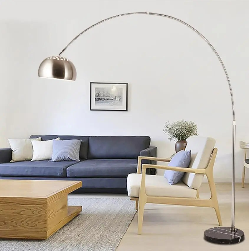 A silver arched floor lamp with a metal dome shade and a black marble base standing over a living room chair.