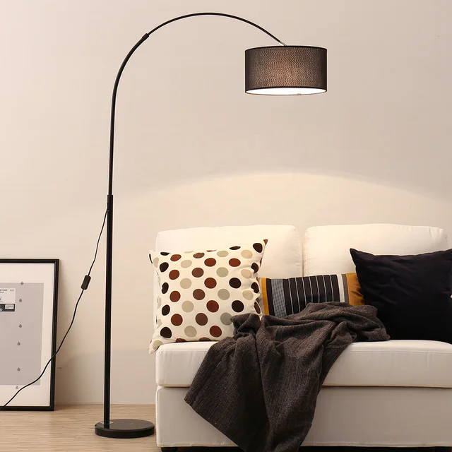 A black arched floor lamp with a dark fabric drum shade standing next to a white sofa with patterned pillows.