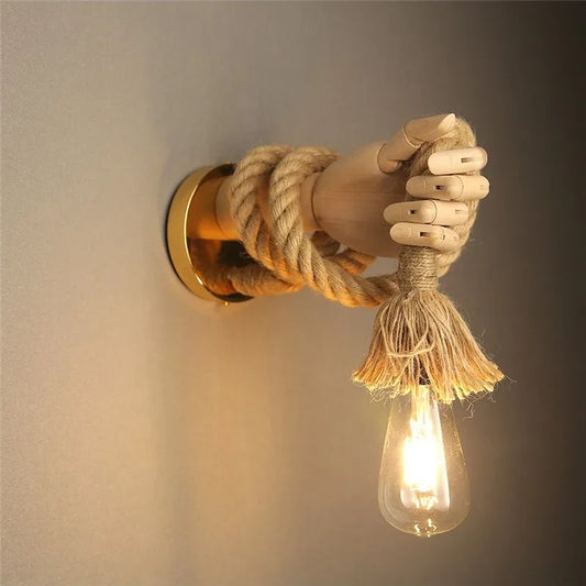A wall-mounted wooden mannequin hand holding a hemp rope with a glowing Edison bulb hanging from the palm.