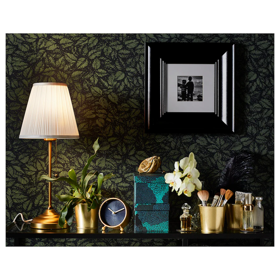 ​Brass table lamp with a white pleated fabric shade and pull chain switch against a dark floral wallpaper.
