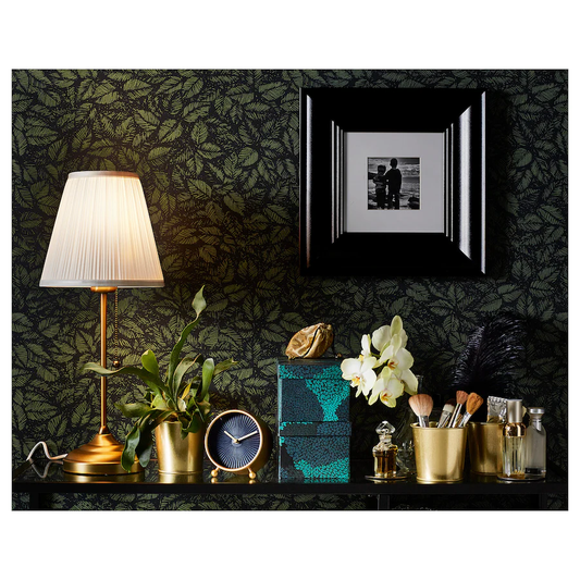 ​Brass table lamp with a white pleated fabric shade and pull chain switch against a dark floral wallpaper.