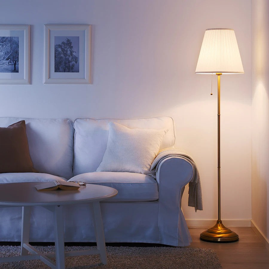 A floor lamp with a cream pleated shade and a brass pole stands next to a white sofa in a styled living room.