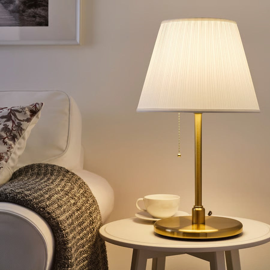 A table lamp with a pleated white shade and a brushed brass base sitting illuminated on a white side table next to a sofa.