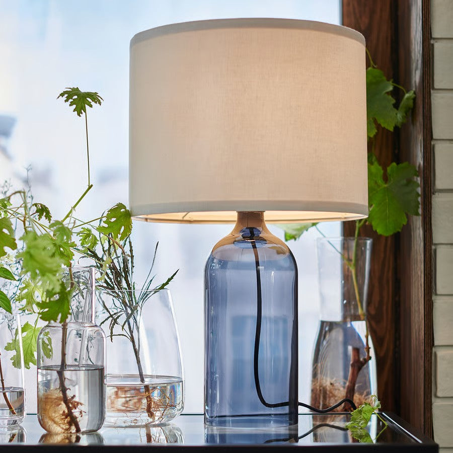 A table lamp with a blue-grey smoke glass base and a white fabric shade sitting on a glass table next to potted plants.
