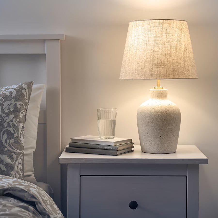A table lamp with a cream speckled ceramic base and a linen-textured lampshade sitting on a white nightstand next to a stack of books.
