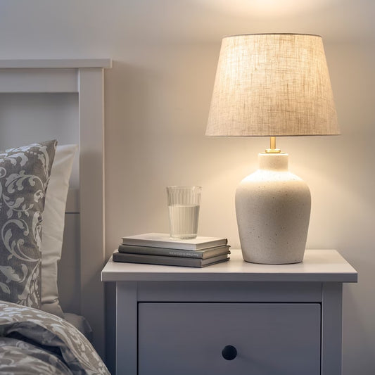 A table lamp with a cream speckled ceramic base and a linen-textured lampshade sitting on a white nightstand next to a stack of books.