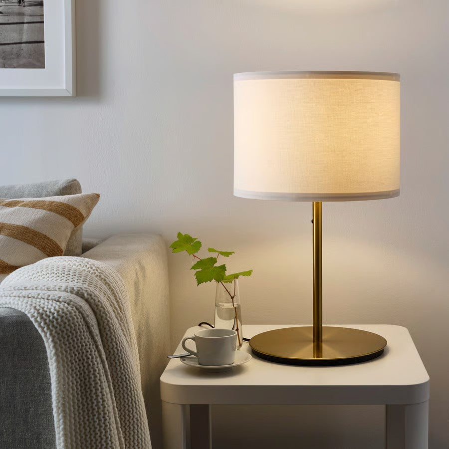 A brass table lamp with a cream fabric drum shade sitting on a white side table next to a cup of coffee.