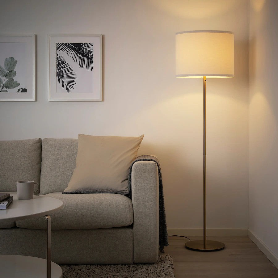 A tall brass floor lamp with a white textile shade standing next to a grey sofa in a living room.