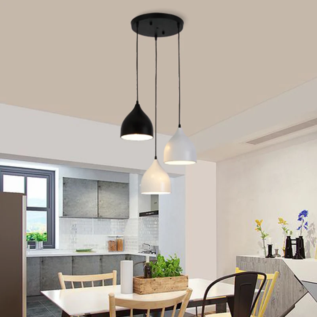 Three modern pendant lights hanging from a ceiling in a bright dining and kitchen area. The lights have black and white dome-shaped shades and are suspended by thin black cords from a round black ceiling mount.