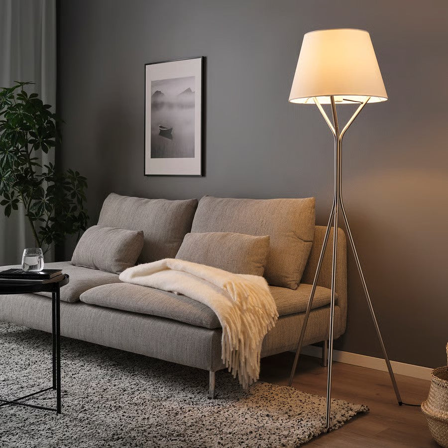 A steel tripod floor lamp with a white fabric shade standing beside a gray couch.