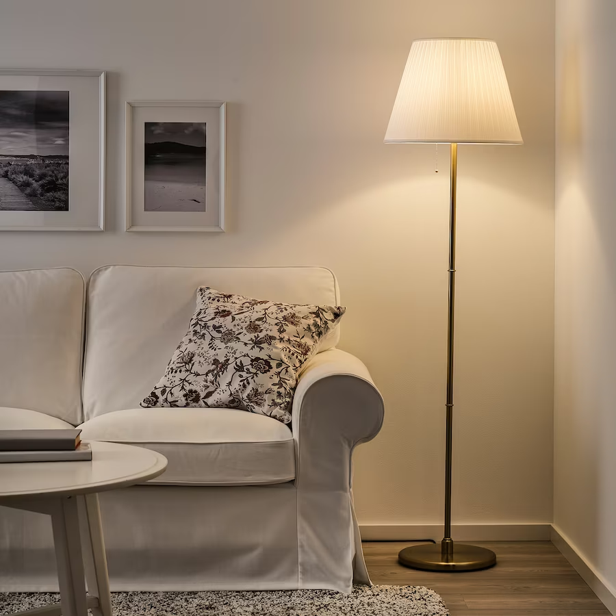 A brass floor lamp with a white pleated shade illuminating a living room corner next to a white sofa as shown in image.png.